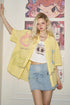 Person wearing a yellow oversized shirt with graphic designs, white graphic t-shirt, and denim skirt in front of a wall with posters.